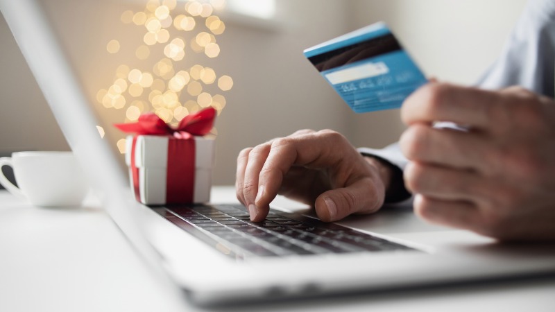 close up of someone holding a credit card in one hand and typing on a laptop in the other. A small gift box is next to the laptop.